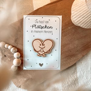 Peut inclure: Épinglette en bois en forme de cœur avec un visage souriant et des bras tenant un biscuit, sur une carte avec le texte "Du hast ein Plätzchen in meinem Herzen". La carte est sur une surface en bois.