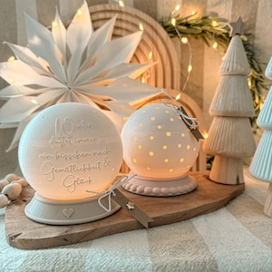 Puede incluir: Dos luces decorativas esféricas de color blanquecino con un brillo cálido, una con texto y la otra con lunares, se encuentran sobre una tabla de madera. Un árbol de Navidad de madera y una estrella de papel están en el fondo. El texto de la primera luz dice "Winter duftet immer ein bisschen nach Gemütlichkeit & Glück."
