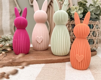May include: Four pastel-coloured bunny figurines in pink, peach, green, and orange, each with a heart detail. The bunnies are displayed on a wooden board. The figurines are approximately 15 cm tall.