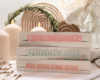 Puede incluir: Tres libros decorativos de madera con texto en rosa y azul claro. El libro superior dice "LIEBLINGSBUCH", el libro del medio dice "SEITENWEISE GLÜCK" y el libro inferior dice "NUR NOCH EINE SEITE". Los libros están sobre una bandeja blanca con un diseño a rayas rosas y blancas.
