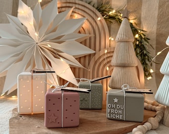 May include: A festive holiday scene featuring decorative gift boxes in pink, green, and cream, adorned with star and dot patterns. A wooden star, tree, and rainbow arch add to the seasonal decor. The boxes have tags and are tied with string.