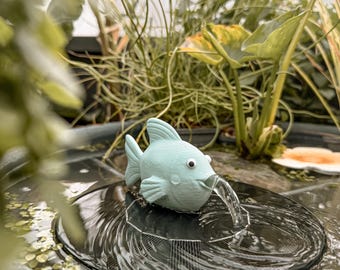 Könnte beinhalten: Ein hellblauer, fischförmiger Springbrunnenaufsatz mit Wackelaugen, montiert auf einem runden, schwarzen Solarpanel, das im Wasser schwimmt. Der Fisch hat eine kleine Öffnung im Maul, aus der ein stetiger Wasserstrahl in das Becken fließt.