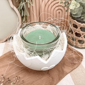 Op de afbeelding: Een groene kaars in een heldere glazen pot, genesteld in een witte, eivormige houder. De kaars staat op een houten plank, omringd door decoratief stro. De kaarshouder is een decoratief item.