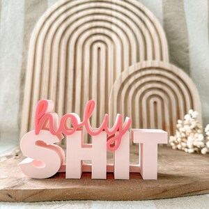 May include: A pink and coral-coloured decorative sign that reads "holy SHIT". The sign is placed in front of two wooden rainbow-shaped arches. The sign is on a wooden surface.