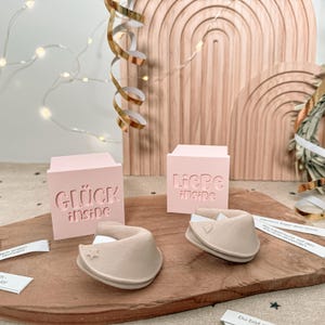 May include: A wooden cutting board displays two pink blocks with the words "GLÜCK INSIDE" and "LIEBE INSIDE" carved into them. Two beige fortune cookie-shaped objects are also on the board, along with small fortune slips.
