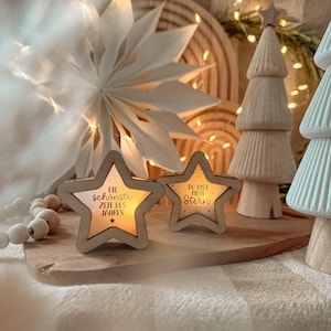 Può includere: Due portacandele a forma di stella in legno con i testi "Die schönste Zeit des Jahres" e "Du bist mein Stern". Le stelle sono su una tavola di legno con altre decorazioni natalizie in legno, tra cui un albero e una stella di carta.