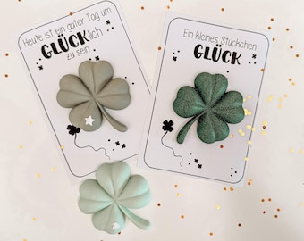 May include: Three-leaf clovers in shades of green and gray, with a white star or heart detail, are displayed on cards with German text. The cards have the word "GLÜCK" and other text. Gold confetti dots the background.