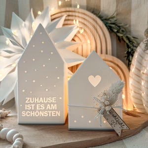 Könnte beinhalten: Zwei weiße Keramik-Kerzenhalter in Hausform mit ausgeschnittenen Punkten und einem Herz. Einer hat den Text "ZUHAUSE IST ES AM SCHÖNSTEN". Der andere hat ein Trockenblumenarrangement und ein Etikett "Wohlfühl Ort".