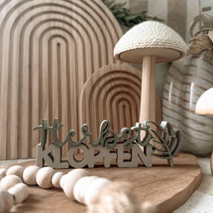 May include: Wooden decorative sign with the text "Herbst Klopfen" in light green and grey. The sign is placed on a wooden cutting board, with a wooden mushroom and other wooden decorative items in the background.