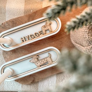 Op de afbeelding: Twee witte en houten kandelaars met decoratieve houten uitsparingen. Eén heeft een Dala-paard, de andere een teckel met een kerstmuts. Het woord "HYGGE" staat op een van de kandelaars.