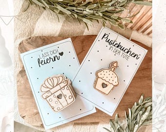 Op de afbeelding: Twee houten kaarten met een lichtblauwe achtergrond en zwarte tekst. De eerste kaart zegt "Lass dich feiern!" en heeft een houten uitsnijding van een cadeaudoos met stippen. De tweede kaart zegt "Pustekuchen in der Tute" en heeft een houten uitsnijding van een cupcake met een kaars.