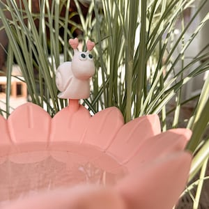 May include: A light pink flower-shaped bird bath with a small, light pink snail figurine attached to the rim. The snail has two antennae with heart-shaped tips and two round black and white eyes. The bird bath has wide, petal-like edges.