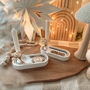Op de afbeelding: Twee witte kandelaars met kaarsen, elk met een klein boeket droogbloemen. Eén houder heeft een houten pompoen, de andere heeft de tekst "Herbst Zauber". Een houten kralenkrans en papieren ster staan op de achtergrond.