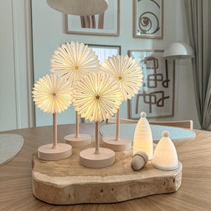 Könnte beinhalten: Vier beleuchtete Papierblumenlampen mit gefalteten weißen Blütenblättern und kleinen goldenen Sternen in der Mitte, jeweils auf einem hellen Ständer. Daneben zwei kleine, leuchtende weiße kegelförmige Keramikornamente und eine Holzeichel auf einer Holzscheibe.