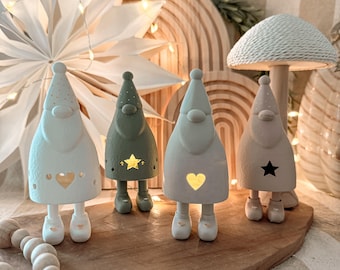 May include: Four ceramic gnome figurines in white, green, and beige, each with a different star or heart cutout. The gnomes have pointed hats and small feet. They are arranged on a wooden board with a wooden mushroom lamp and starburst decoration in the background.