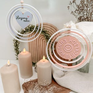 Puede incluir: Dos adornos colgantes decorativos con diseños circulares. Uno presenta un corazón con el texto "Herzens Mensch", el otro tiene una carita sonriente y el texto "Rundum Glücklich". Los adornos son blancos, rosas y azules. Velas beige y elementos decorativos en el fondo.