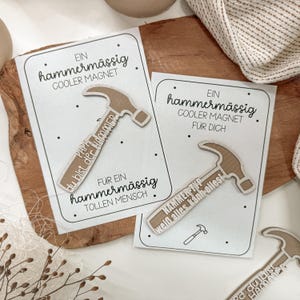 Puede incluir: Dos imanes beige con forma de martillo sobre tarjetas blancas. Las tarjetas tienen texto negro que dice "Ein hammermässig COOLER MAGNET" y "FÜR DICH". Un imán dice "PAPA du bist der HAMMER" y el otro "HAMMER-TYP weiß alles kann-alles!"