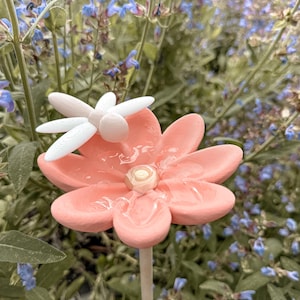 Könnte beinhalten: Ein Gartenstecker mit einer matten, pfirsichfarbenen Blume, die ein glänzendes, durchscheinendes Zentrum hat und auf deren Blütenblatt eine weiße Libelle sitzt. Die Blume ist auf einem dünnen, hellen Stab befestigt und dient als Dekoration.