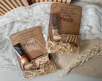 May include: Two brown wooden signs with the text "In meiner Hand Schokolade In meinem Herzen Dankbarkeit Merci" and a chocolate bar on each. The signs are surrounded by shredded paper.
