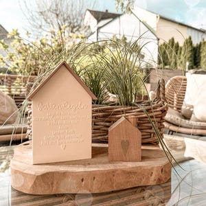 Può includere: Due decorazioni in legno a forma di casa. Una è di colore beige chiaro con il testo "Garten Regeln" e altro testo. L'altra è una piccola casa in legno con un cuore intagliato. Entrambe sono su una lastra di legno.