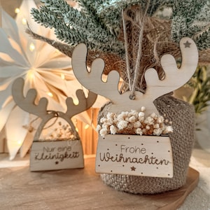 Puede incluir: Adornos navideños de reno de madera con cuernos, rellenos de flores blancas secas. Un adorno tiene el texto "Frohe Weihnachten" y el otro dice "Nur eine Kleinigkeit". Ambos cuelgan de hilo.