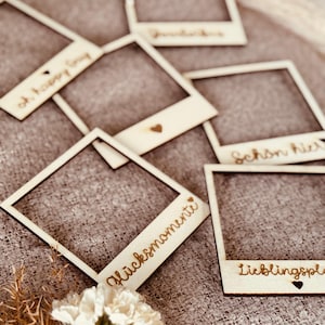 Könnte beinhalten: Holzrahmen im Polaroid-Stil mit verschiedenen Schriftzügen und Herz-Akzenten. Die Rahmen sind auf einer strukturierten Oberfläche angeordnet, mit einigen weißen Blumen im Vordergrund. Sätze wie "Glücksmomente" sind zu sehen.
