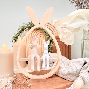 May include: An Easter-themed decorative shelf in the shape of an egg with bunny ears, painted in a light peach colour. Two bunny figurines, one peach and one white, sit on a wooden shelf. The background includes candles and decorative elements.