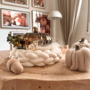 Puede incluir: Un recipiente decorativo de cristal ámbar con forma de calabaza se asienta dentro de un anillo trenzado blanco, acompañado de una pequeña bellota de madera y una calabaza de tela beige. La calabaza de cristal tiene un acabado metálico.