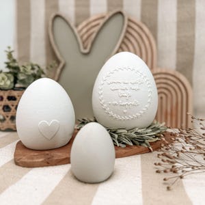 Puede incluir: Tres huevos de Pascua decorativos blancos de diferentes tamaños, uno con un corazón, otro con texto dentro de una corona y otro liso. Los huevos están sobre una tabla de madera con una silueta de conejo y otras decoraciones de Pascua en el fondo.