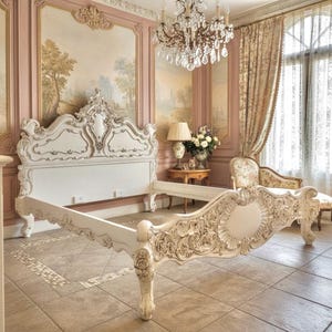 May include: An ornate, white bed frame with detailed carvings. The headboard features a decorative crest, and the footboard has elaborate floral and scrollwork. The bed frame is set in a room with a chandelier and floral wall decor.