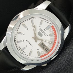 May include: A Seiko 5 automatic wristwatch with a white dial, silver-tone hour markers, and a black leather strap. The dial features a red accent segment near the three o'clock position, a day-date window, and the text Seiko, Automatic, and Japan Made.