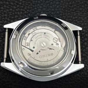 Puede incluir: Vista de cerca de la parte trasera de un reloj Seiko plateado, mostrando el intrincado mecanismo interno. El movimiento del reloj es visible a trav&eacute;s de un cristal transparente, con las palabras "Japan Seventeen 17 Jewels" y "Seiko".