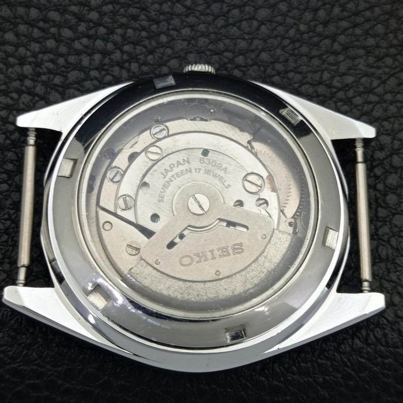 Puede incluir: Vista de cerca de la parte trasera de un reloj Seiko plateado, mostrando el intrincado mecanismo interno. El movimiento del reloj es visible a trav&eacute;s de un cristal transparente, con las palabras "Japan Seventeen 17 Jewels" y "Seiko".