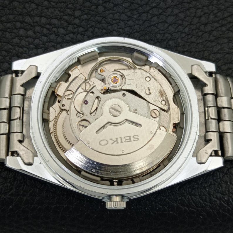 May include: A silver-toned Seiko watch with a visible mechanical movement. The watch's back is open, revealing the intricate internal gears, springs, and the word "SEIKO" on a silver plate. The watch band is also silver-toned.