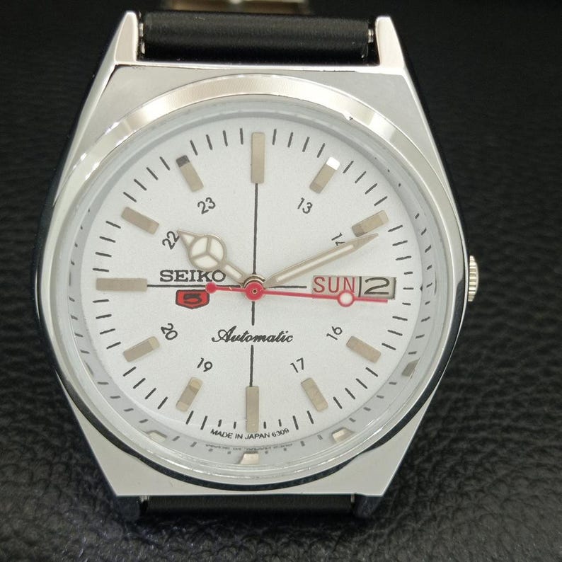 May include: A silver Seiko 5 automatic watch with a white face. The watch features a black leather band, a day and date display, and luminous hands and markers. The watch face displays the text "SEIKO 5 Automatic" and "MADE IN JAPAN 6309".