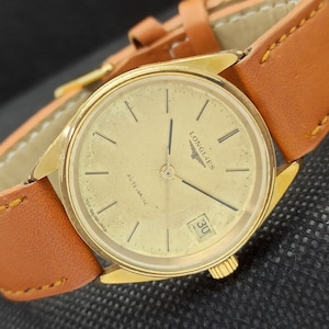 May include: A gold-tone Longines watch with a white face and black hands. The watch has a date window at the 3 o'clock position and the words "Longines" and "Automatic" printed on the face.