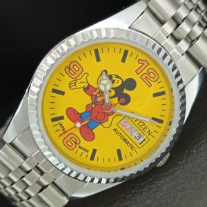 Op de afbeelding: Een vintage Citizen automatisch polshorloge met een gele wijzerplaat met een stripfiguur. Het horloge heeft een zilverkleurige metalen band en een datumweergave. De wijzerplaat heeft zwarte markeringen en de tekst "JAPAN MADE".