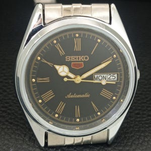 May include: A silver-toned Seiko 5 automatic wristwatch with a black dial. The watch features gold-colored hands and Roman numeral hour markers. The day and date are displayed at the 3 o'clock position. The watch band is silver-toned.