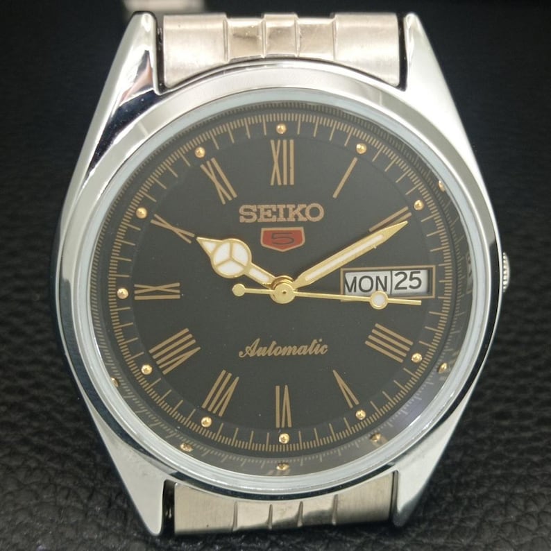 May include: A silver-toned Seiko 5 automatic wristwatch with a black dial. The watch features gold-colored hands and Roman numeral hour markers. The day and date are displayed at the 3 o'clock position. The watch band is silver-toned.