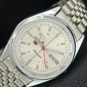 May include: A silver-tone Seiko 5 automatic wristwatch with a cream-colored dial, featuring a red second hand, a day-date window at the three o'clock position, and a stainless steel link bracelet. The dial displays the text Seiko, 5, and Automatic.