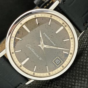 Puede incluir: Primer plano de un reloj vintage Eterna-Matic Centenaire 60. El reloj tiene una caja plateada, una esfera gris oscura con agujas e indicadores plateados y una ventana de fecha. El reloj tiene una correa de cuero negro.