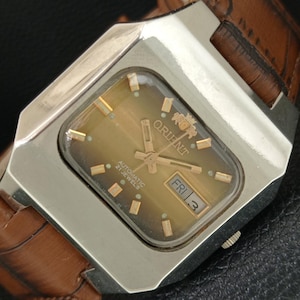 May include: A vintage Orient automatic wristwatch with a rectangular silver-tone case and a brown leather strap. The dial is a gradient tan color featuring gold-tone hour markers, gold hands, and a day-date window displaying FRI 3.