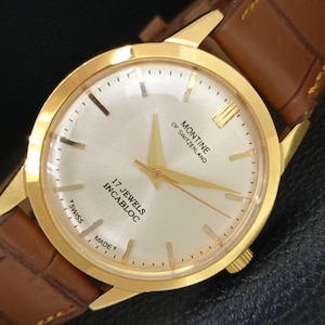 K&ouml;nnte beinhalten: Eine goldfarbene Armbanduhr mit einem runden, geb&uuml;rsteten silbernen Zifferblatt, goldenen Zeigern und goldenen Stundenmarkierungen. Das Zifferblatt tr&auml;gt den Text MONTINE OF SWITZERLAND, 17 JEWELS INCABLOC und T SWISS MADE T. Sie hat ein braunes Lederarmband.