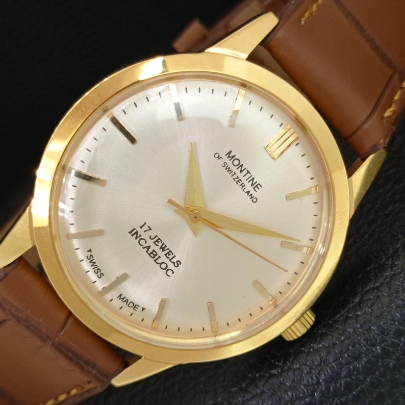 K&ouml;nnte beinhalten: Eine goldfarbene Armbanduhr mit einem runden, geb&uuml;rsteten silbernen Zifferblatt, goldenen Zeigern und goldenen Stundenmarkierungen. Das Zifferblatt tr&auml;gt den Text MONTINE OF SWITZERLAND, 17 JEWELS INCABLOC und T SWISS MADE T. Sie hat ein braunes Lederarmband.