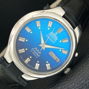 May include: A silver-tone wristwatch with a vibrant blue gradient dial, featuring silver hour markers, luminous hands, and a day-date window at the three o'clock position. The dial displays the text Orient, AAA Crystal, and 21 Jewels.