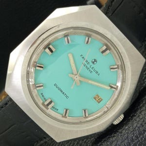 May include: A silver watch with a turquoise face and black markings. The watch face has the text "DUOMATIC" and "FAIVRE-LEUBA GENEVE" and a date window showing the number 26.