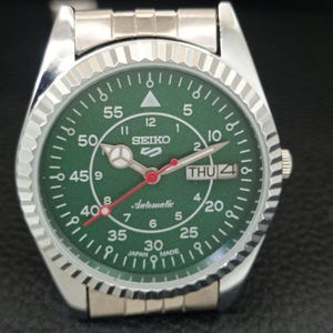 May include: Silver-toned Seiko 5 automatic watch with a green dial. The watch features a fluted bezel, a day-date display, and a red second hand. The dial includes white numerals and markers, with the word "Automatic" printed in cursive.