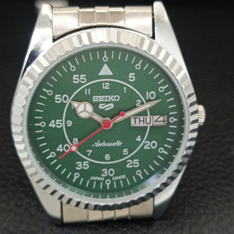 May include: Silver-toned Seiko 5 automatic watch with a green dial. The watch features a fluted bezel, a day-date display, and a red second hand. The dial includes white numerals and markers, with the word "Automatic" printed in cursive.