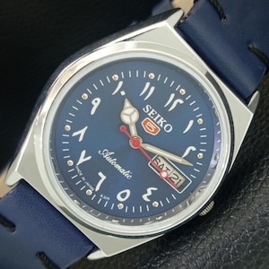 May include: A Seiko 5 automatic wristwatch with a round silver-tone stainless steel case and a blue dial. The dial features Arabic-Indic numerals, a day-date window at the three o'clock position, and a red second hand. It has a blue leather strap.