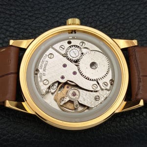 K&ouml;nnte beinhalten: Eine goldfarbene Armbanduhr mit offenem mechanischem Uhrwerk, das durch den Geh&auml;useboden sichtbar ist. Das Werk zeigt silberfarbene Zahnr&auml;der, eine Unruh und die Aufschrift SWISS SEVENTEEN 17 JEWELS. Mit braunem Lederarmband.